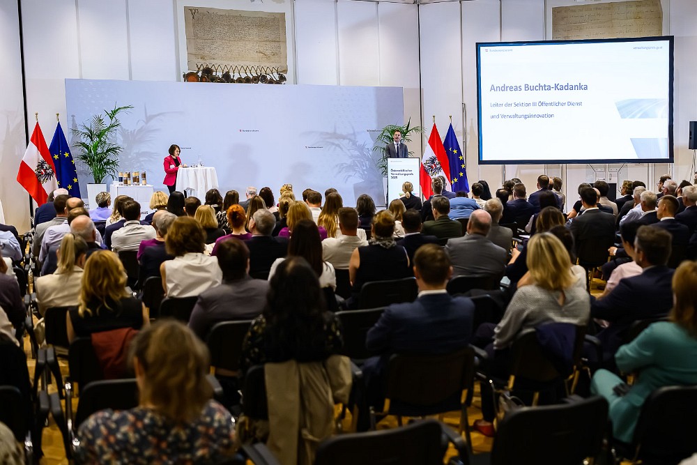 Eröffnungsveranstaltung des Österreichischen Verwaltungspreises 2025. Eine Rednerin spricht auf der Bühne vor vollbesetztem Saal. Im Hintergrund zwei Flaggen (Österreich und EU), eine Leinwand mit der Begrüßungsfolie und Pokale auf einem Stehtisch.