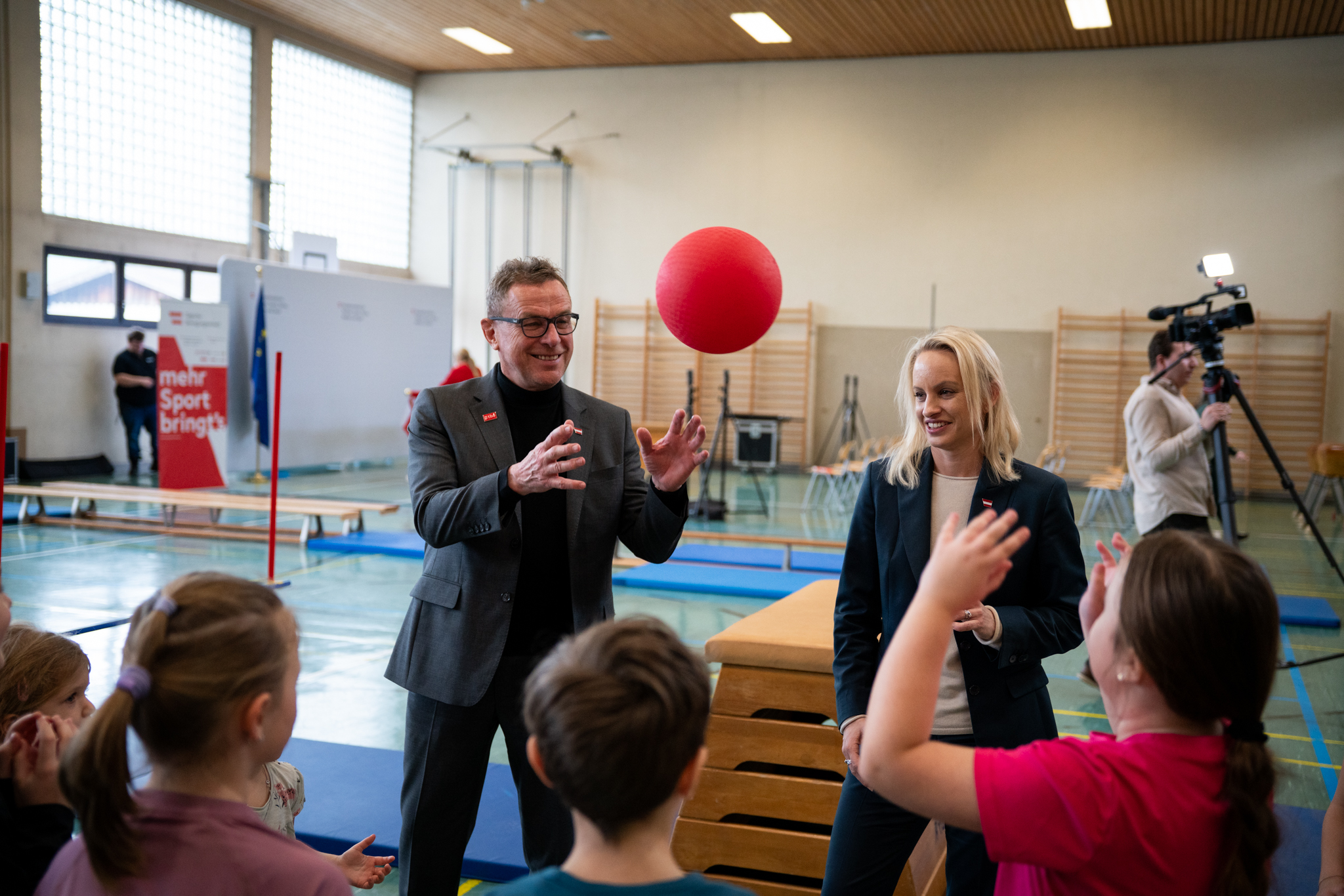 Ralf Rangnick und Sport-Staatssekretärin Michaela Schmidt stehen in einer Turnhalle vor Kindern; ein roter Ball fliegt zwischen ihnen.