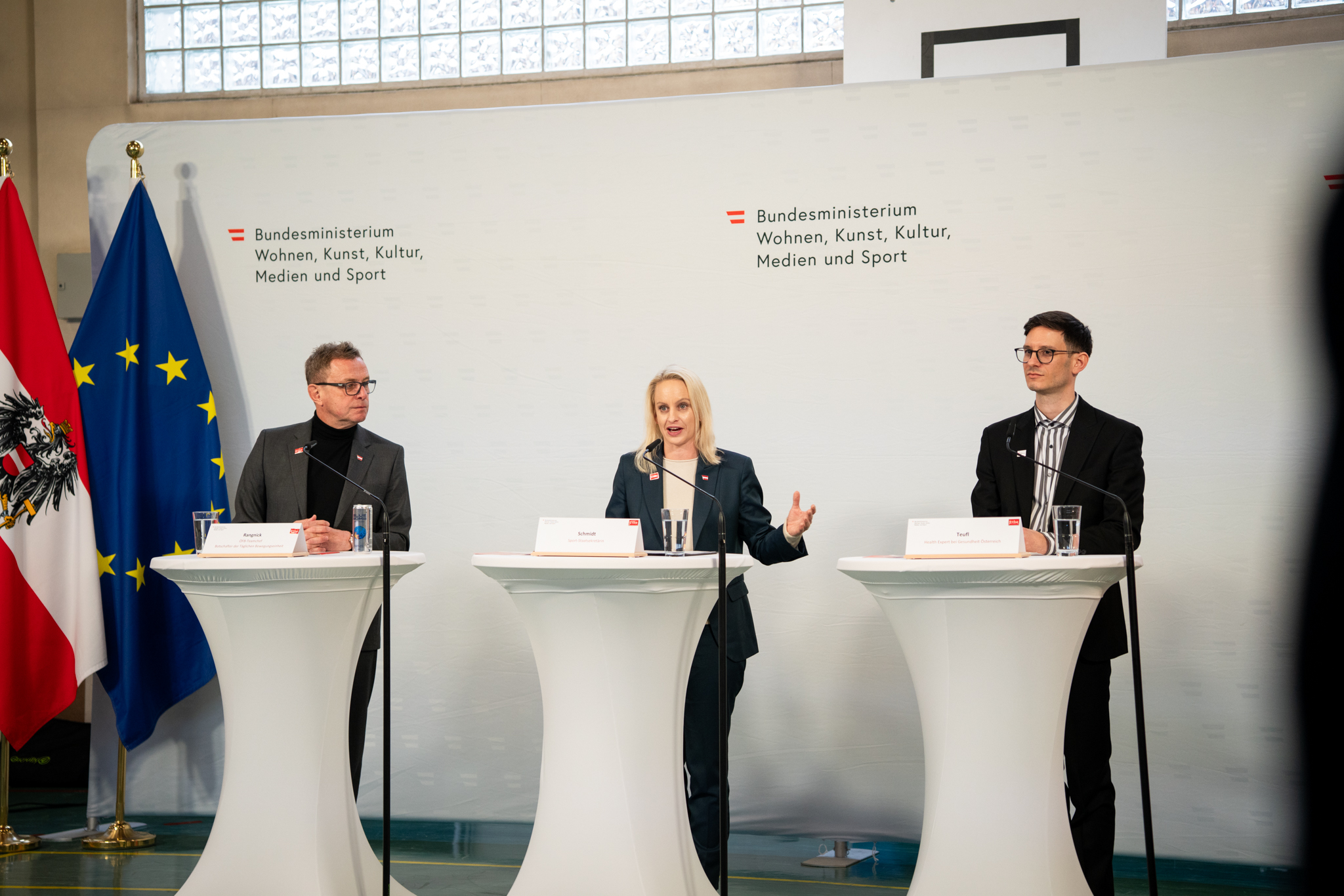 Pressegespräch zur Täglichen Bewegungseinheit: Ralf Rangnick, Sport-Staatssekretärin Michaela Schmidt und Lukas Teufl stehen an Stehtischen vor Österreich- und EU-Flagge.