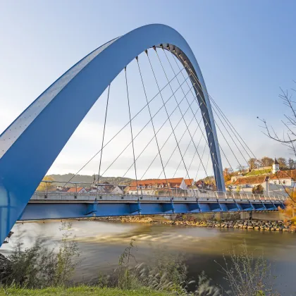 Moderne blaue Bogenbrücke über die Mur in Bruck an der Mur, mit Blick auf Altstadt und Kirche am sonnigen Herbsttag.