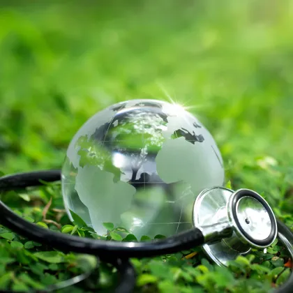 Eine gläserne Weltkugel liegt auf einer grünen Wiese und wird von einem Stethoskop umgeben. Das Motiv symbolisiert die Verbindung von Gesundheit, Medizin und globaler Nachhaltigkeit – im Sinne einer umweltbewussten, ressourcenschonenden Gesundheitsversorgung.
