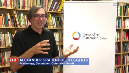 Mag. Alexander Grabenhofer-Eggerth, Abteilungsleiter für Psychosoziale Gesundheit, spricht in einem Interview, im Hintergrund Bücherregal und ein Banner mit dem Logo der Gesundheit Österreich GmbH.