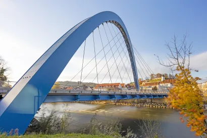 Moderne blaue Bogenbrücke über die Mur in Bruck an der Mur, mit Blick auf Altstadt und Kirche am sonnigen Herbsttag.