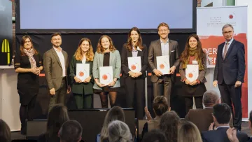 Sieben Personen stehen auf einer Bühne, einige halten Urkunden, ein Publikum applaudiert.