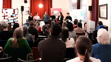 Ein Publikum hört einer Diskussion mit fünf Sprecher:innen zu, im Hintergrund ein AIDS-Hilfe-Wien-Banner.