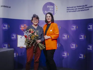 Zwei Frauen vor einem Pride Biz Austria Banner, eine hält Blumen und eine Urkunde.