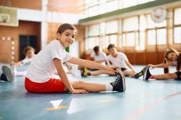 Ein Kind in Sportkleidung dehnt sich in einer Turnhalle, während andere Kinder ebenfalls Dehnübungen machen.