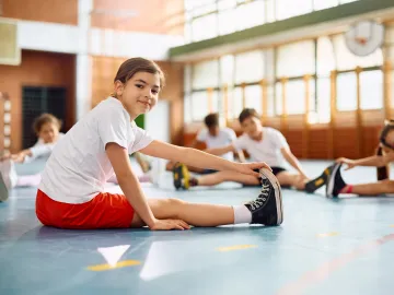 Ein Kind in Sportkleidung dehnt sich in einer Turnhalle, während andere Kinder ebenfalls Dehnübungen machen.