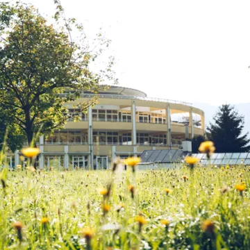 Beispiel klimafreundliche Gesundheitseinrichtung mit großer Fensterfassade, umgeben von Bäumen und einer blühenden Wiese – Symbol für Nachhaltigkeit im Gesundheitswesen.