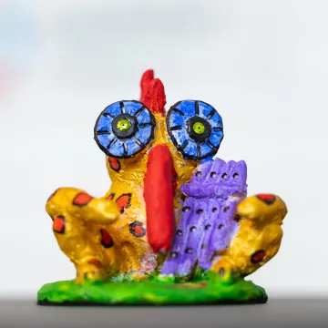 Buntes, handbemaltes Papageno-Figur-Modell mit übergroßen Augen und roten Flecken, auf grünem Sockel vor weißem Hintergrund.