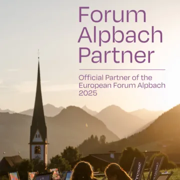 Panorama von Alpbach bei Sonnenuntergang mit dem markanten Kirchturm im Vordergrund und den Tiroler Bergen im Hintergrund. Rechts im Bild der Schriftzug „Forum Alpbach Partner – European Forum Alpbach"
