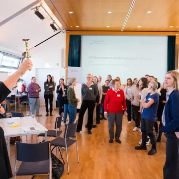 Teilnehmende des Bürger:innenforums zur Stärkung der Patient:innen- und Bevölkerungsbeteiligung im Gesundheitswesen in Wien, organisiert von der Gesundheit Österreich GmbH. Eine Moderatorin eröffnet die Veranstaltung mit einem symbolischen Signalhorn.