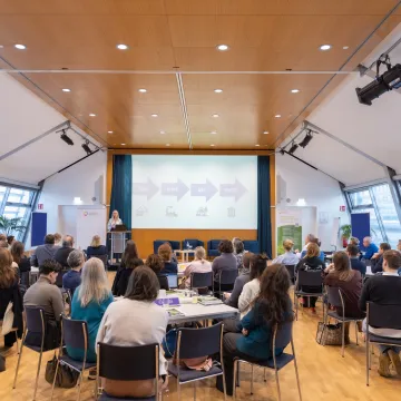 Teilnehmende sitzen im Konferenzsaal und verfolgen einen Vortrag zur Kreislaufwirtschaft im Gesundheitswesen auf der Bühne.