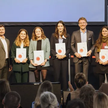© Marlene Fröhlich | luxundlumen Sieben Personen stehen auf einer Bühne, einige halten Urkunden, ein Publikum applaudiert.