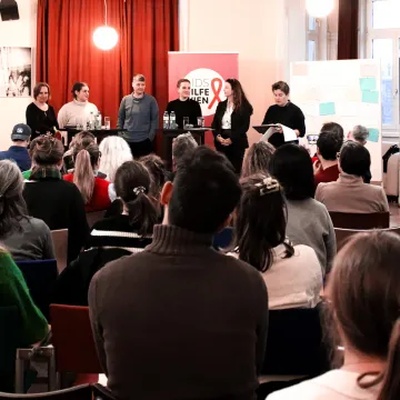 © Aids Hilfe Wien/Klaus Hager Ein Publikum hört einer Diskussion mit fünf Sprecher:innen zu, im Hintergrund ein AIDS-Hilfe-Wien-Banner.