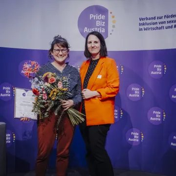 Zwei Frauen vor einem Pride Biz Austria Banner, eine hält Blumen und eine Urkunde.