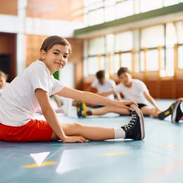 Ein Kind in Sportkleidung dehnt sich in einer Turnhalle, während andere Kinder ebenfalls Dehnübungen machen.