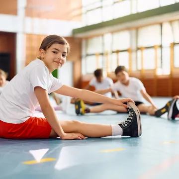 © Drazen – stock.adobe.com Ein Kind in Sportkleidung dehnt sich in einer Turnhalle, während andere Kinder ebenfalls Dehnübungen machen.