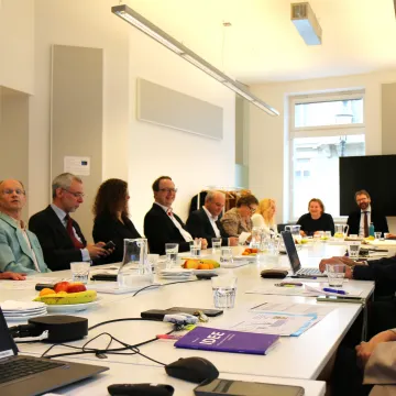 © GÖG / Georg Gatnar Ministerin Korinna Schumann sitzt gemeinsam mit Vertreter:innen der Gesundheit Österreich GmbH an einem langen Konferenztisch. Die Teilnehmenden blicken lächelnd in Richtung Präsentation, auf dem Tisch liegen Unterlagen und Obst.