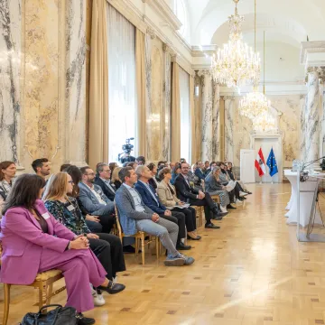 Podiumsdiskussion beim Best Practice Award „Klimafreundliche Gesundheitseinrichtungen“ 2026. Fünf Personen diskutieren an Stehtischen vor einem sitzenden Publikum im Festsaal des Gesundheitsministeriums. Eine Teilnehmerin spricht ins Mikrofon, die Diskussion wird gefilmt.
