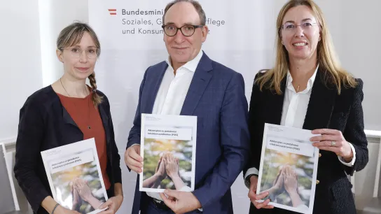 © BKA Regina Aigner Drei Personen halten einen Bericht zu postakuten Infektionssyndromen (PAS) vor einem Ministeriums-Banner.