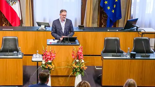 © ORF III Ein Mann spricht an einem Rednerpult, dahinter EU- und Österreich-Flagge.