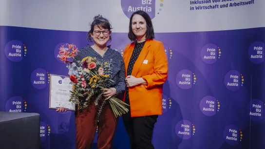 © Christine Miess Zwei Frauen vor einem Pride Biz Austria Banner, eine hält Blumen und eine Urkunde.