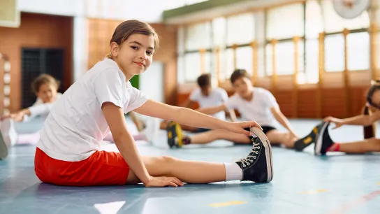 Ein Kind in Sportkleidung dehnt sich in einer Turnhalle, während andere Kinder ebenfalls Dehnübungen machen.