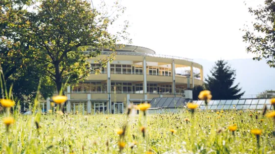 © Christine Kostner Beispiel klimafreundliche Gesundheitseinrichtung mit großer Fensterfassade, umgeben von Bäumen und einer blühenden Wiese – Symbol für Nachhaltigkeit im Gesundheitswesen.