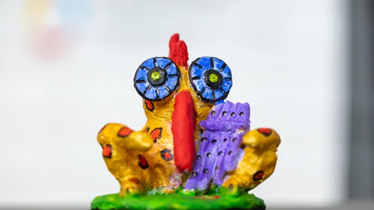 © BKA / Valentin Brauneis Buntes, handbemaltes Papageno-Figur-Modell mit übergroßen Augen und roten Flecken, auf grünem Sockel vor weißem Hintergrund.