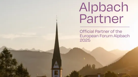 Panorama von Alpbach bei Sonnenuntergang mit dem markanten Kirchturm im Vordergrund und den Tiroler Bergen im Hintergrund. Rechts im Bild der Schriftzug „Forum Alpbach Partner – European Forum Alpbach"