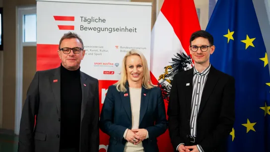 © BMWKMS/Schlögl Ralf Rangnick, Sport-Staatssekretärin Michaela Schmidt und Lukas Teufl stehen bei einem Pressetermin zur „Täglichen Bewegungseinheit“ vor einem Roll-up sowie Österreich- und EU-Flaggen.